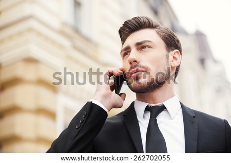 Staying in touch. Low angle view of confident young man in formalwear holding mobile phone and looking away while standing outdoors