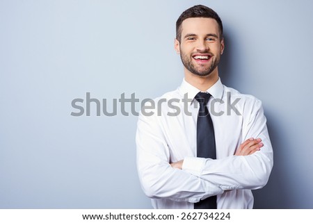Young and successful. Portrait of handsome young man in shirt and tie keeping arms crossed and smiling while standing against grey background