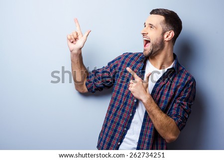 Just look at that! Happy young man in casual shirt pointing away and smiling while standing against grey background