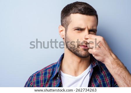 Disgusting smell. Portrait of frustrated young man in casual shirt holding nose and expressing negativity while standing against grey background