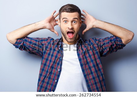 This is amazing! Surprised young man in casual shirt keeping mouth open and staring at camera while standing against grey background
