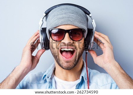 Enjoying his favorite song. Happy young stylish man in headphones expressing positivity and looking at camera while standing against grey background