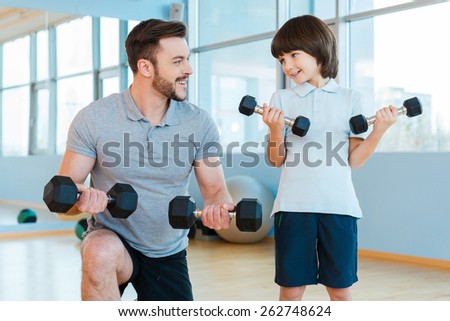 Exercising together. Happy father and son exercising with dumbbells and smiling while standing in health club