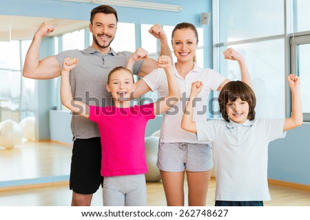 Proud to be strong and healthy. Happy sporty family showing their biceps and smiling while standing close to each other in sports club