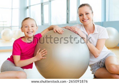 Girls in sports club. Cheerful mother and daughter leaning at the fitness ball and smiling while sitting on the floor in sports club