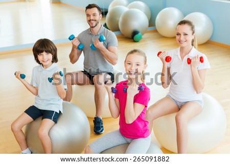 Enjoying time in sorts club. Top view of happy sporty family exercising with dumbbells in sports club while sitting on the fitness balls together