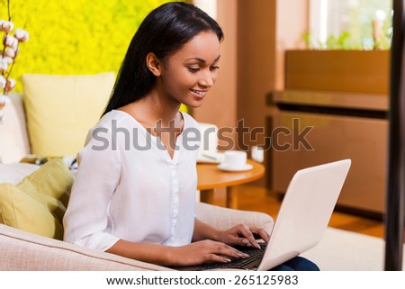 She loves working at home. Attractive young African woman working on laptop and smiling while sitting at the chair at home