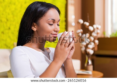 Enjoying fresh coffee. Side view of beautiful young African woman holding coffee cup near nose and smiling while keeping eyes closed and sitting at the chair at home