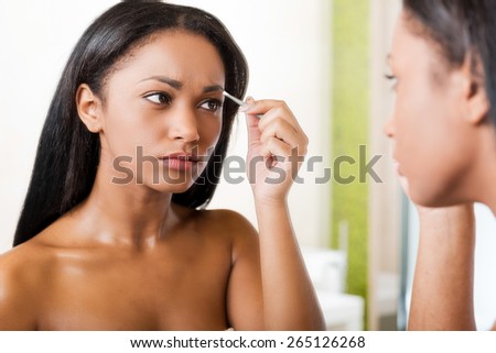 Woman tweezing eyebrows. Concentrated young African woman tweezing her eyebrows and looking at herself in a mirror