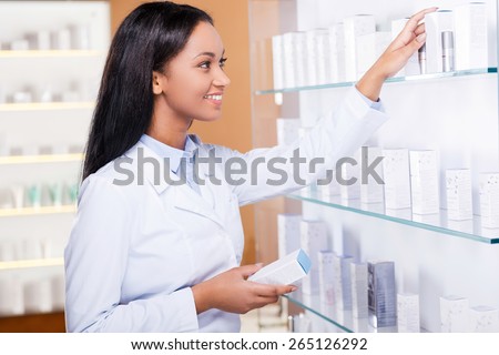 Choosing the right medicine for you. Beautiful young African woman in lab coat choosing medicine while standing near the self in drugstore