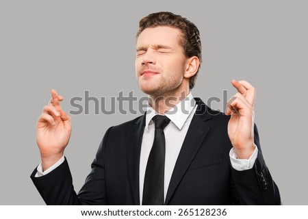 Waiting for special moment. Portrait of young man in formalwear keeping fingers crossed and eyes closed while standing against grey background