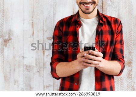 His smart helper. Cropped picture of handsome young man holding mobile phone and smiling while standing against the wooden wall