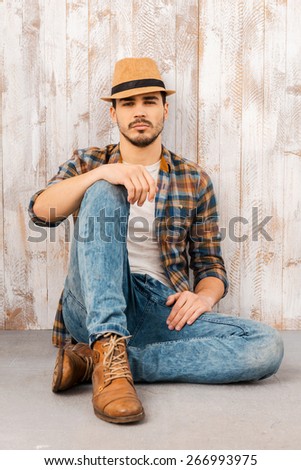 Calm and confident. Handsome young man wearing hat and looking at camera while sitting against the wooden wall