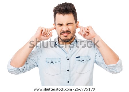 This is too loud! Frustrated young man in shirt holding fingers in his ears and keeping eyes closed while standing against white background