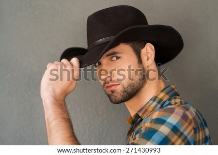 Wild West is his heart. Handsome young man adjusting his cowboy hat and looking at camera while standing against grey background