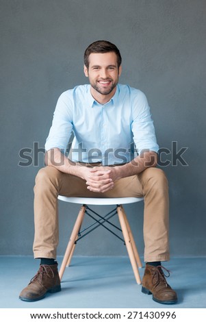 Positive thinker. Full length of handsome young man looking at camera and smiling while sitting against grey background