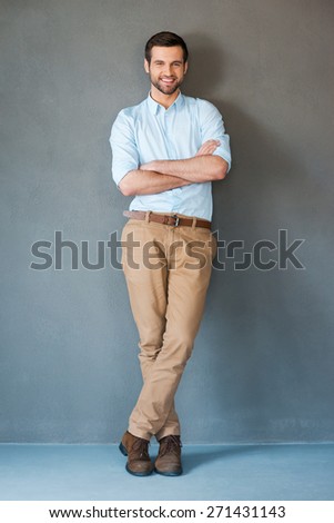 Only positive emotions. Full length of handsome young man in shirt keeping arms crossed and smiling at camera while standing against grey background