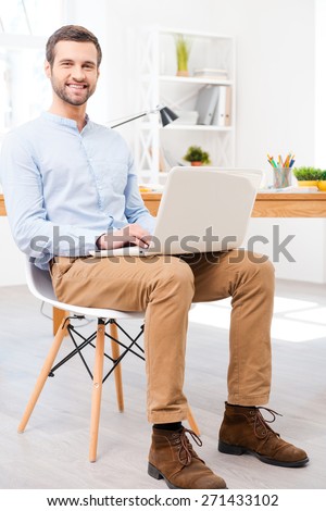 Easy to work. Handsome young man in shirt working on laptop in front of his desk and smiling at camera while sitting at office
