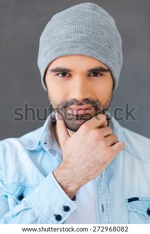 He wants to melt your heart. Portrait of handsome young man wearing hat and looking at camera while standing against grey background