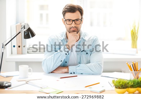Generating new ideas. Handsome young man in shirt and eyewear holding hand on chin and looking at camera while sitting at his working place