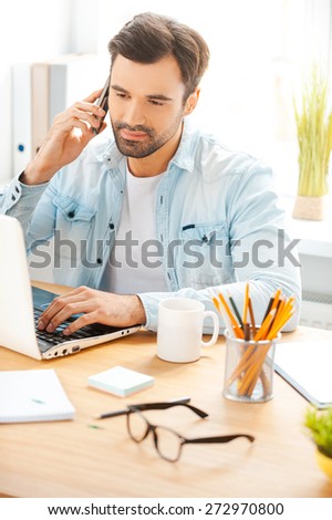 He is always hard at work. Handsome young man in shirt talking on the telephone and working on laptop while sitting at his working place