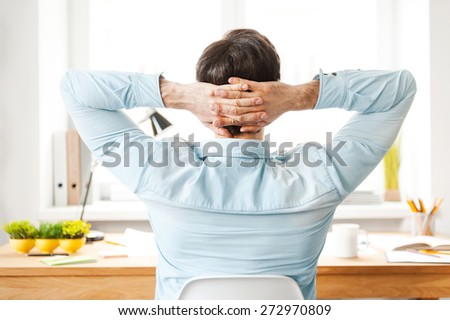 Having rest for productive work. Rear view of young man in shirt holding head in hands while sitting at his working place