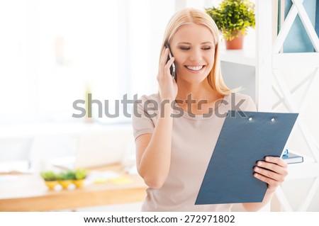 Discussing new project. Beautiful young woman leaning at the shelf in office and smiling while talking on the mobile phone and looking at clipboard