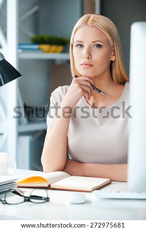 Confident business expert. Beautiful young woman holding hand on chin and looking at camera while sitting at her working place in office