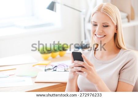 Good news from colleagues. Beautiful young woman holding mobile phone and smiling while sitting at her working place in office