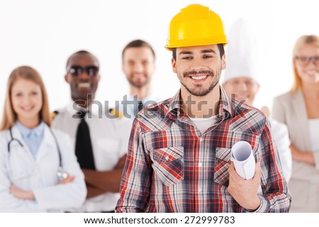 When I grow up I will be an engineer. Confident young man in hardhat holding blueprint and smiling while group of people in different professions standing in the background