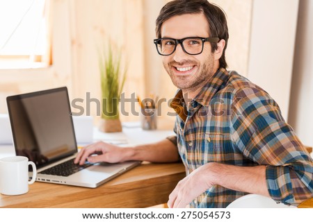 Great mood for working good. Handsome young man working on his laptop and looking at camera over shoulders while sitting at his working place
