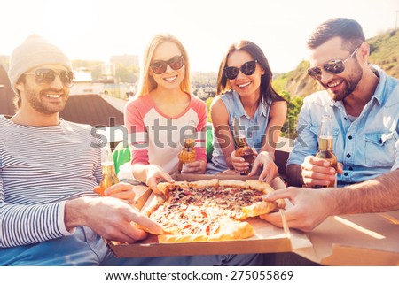 Time for pizza! Four young cheerful people eating pizza and drinking beer while sitting at the bean bags on the roof of the building