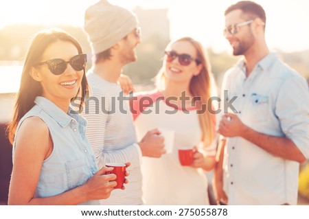 Enjoying coffee break. Beautiful young woman holding coffee cup and smiling while standing near her friends on the roof terrace