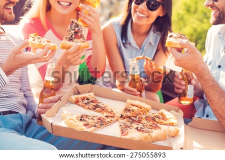 Pizza time! Close-up of four young cheerful people eating pizza and drinking beer outdoors