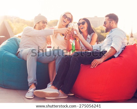Cheers to us! Four young cheerful people cheering with beer and smiling while sitting at the bean bags on the roof of the building