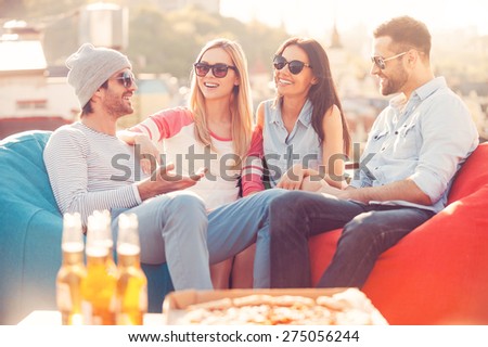 Spending great time together. Four young cheerful people chatting while sitting at the bean bags on the roof of the building with pizza and beer laying on foreground