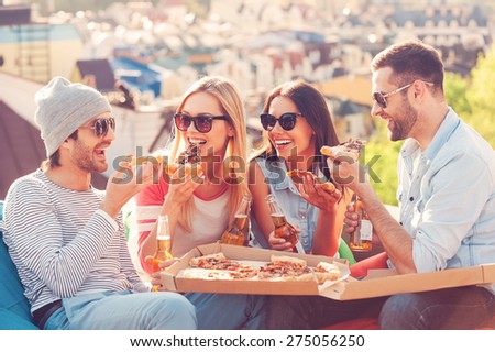 Pizza time. Four young happy people eating pizza and drinking beer while sitting at the bean bags on the roof of the building