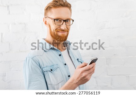 Confident smile and perfect style. Handsome young bearded man holding mobile phone and smiling at camera while standing against brick wall