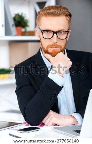 Leading this company. Concentrated bearded businessman and holding hand on chin and looking at camera while sitting at his working place
