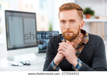 Office worker at his desk. Thoughtful young bearded man keeping hands clasped and looking at camera while sitting at his working place
