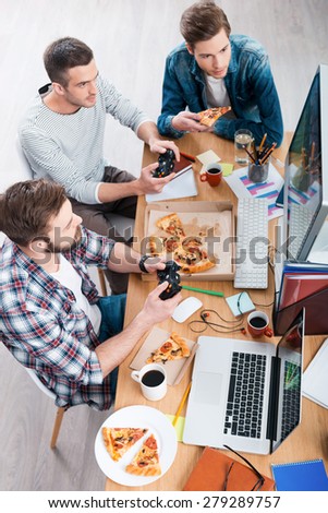 Relaxing after work. Top view of three young men playing computer games and eating pizza while sitting at the desk