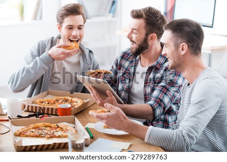 Pizza time! Three young cheerful men eating pizza together while sitting in the office