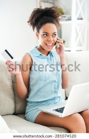 Enjoying online shopping. Attractive young African woman talking on the mobile phone and holding credit card while sitting on the couch with laptop at her knees