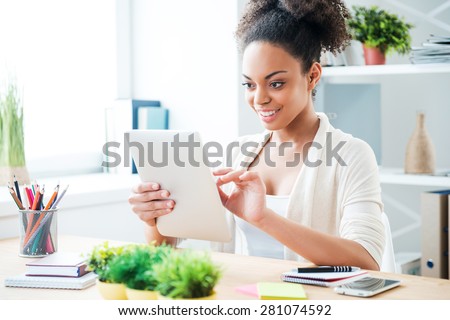 Working on tablet. Beautiful young African woman working on digital tablet and smiling while sitting at her working place in office