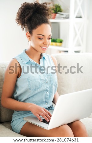 Beauty with laptop. Attractive young African woman working on laptop and smiling while sitting on the couch at home