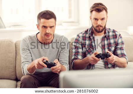 Focused on game. Two concentrated young men playing video games while sitting on sofa