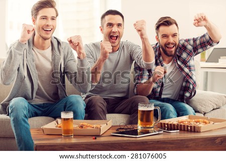 Domestic fans. Three happy young men watching football game and keeping arms raised while sitting on sofa