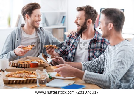 We work hard but have fun doing it! Three happy young men eating pizza together while sitting in the office