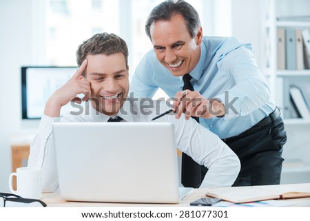 Lending him a hand with his work. Two cheerful business people in formalwear discussing something and looking at laptop