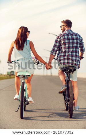 Perfect date. Rear view of young couple holding hands while riding on bicycles along the road